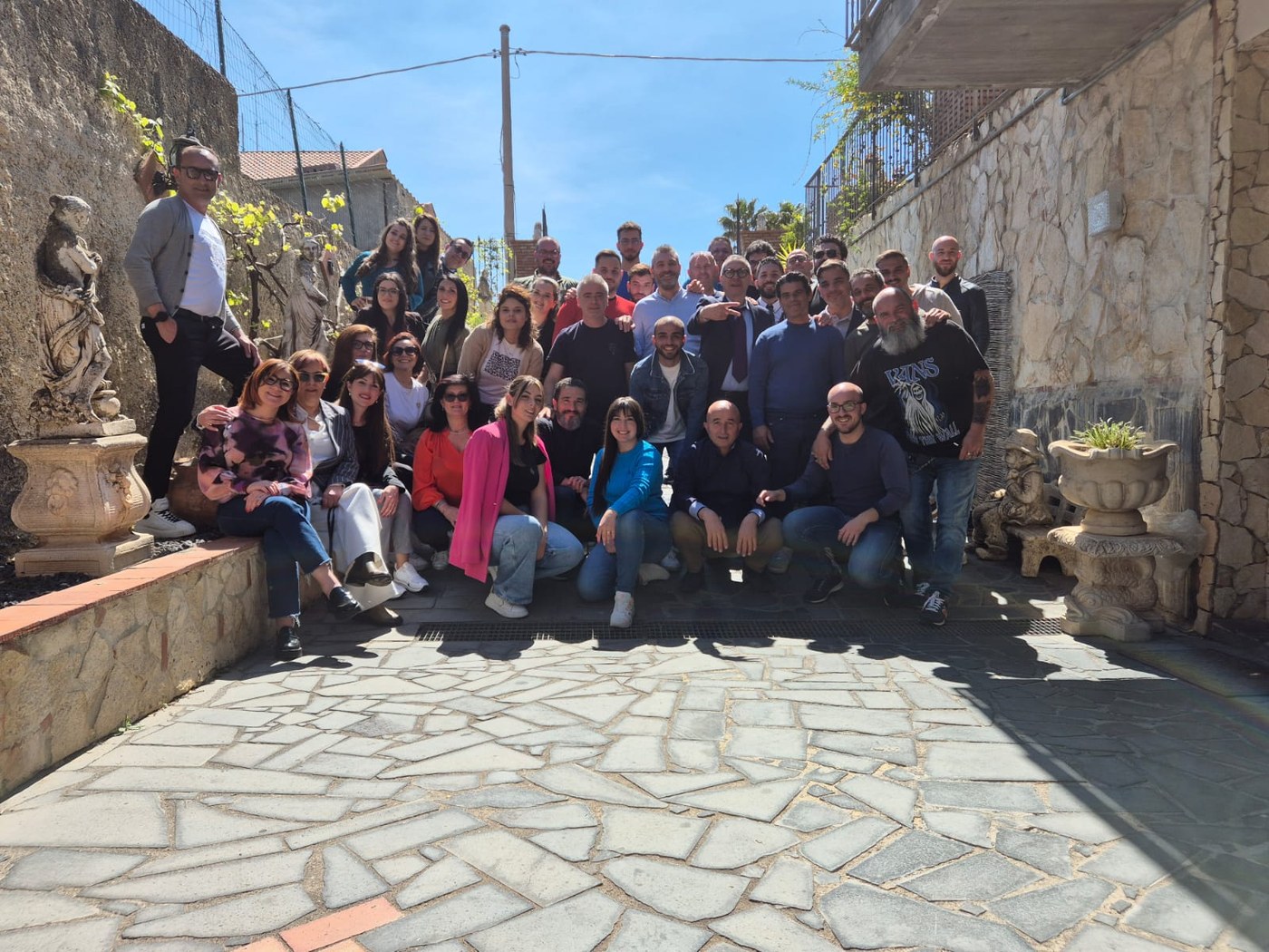 Gruppo di circa quaranta persone sorridenti in posa su un cortile lastricato in pietra, tra muri rocciosi e statue