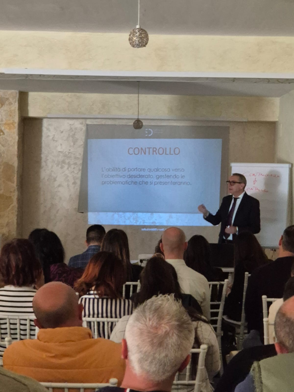 Relatore in completo scuro davanti alla slide «CONTROLLO» con logo Soluzioni Differenti, pubblico su sedie bianche
