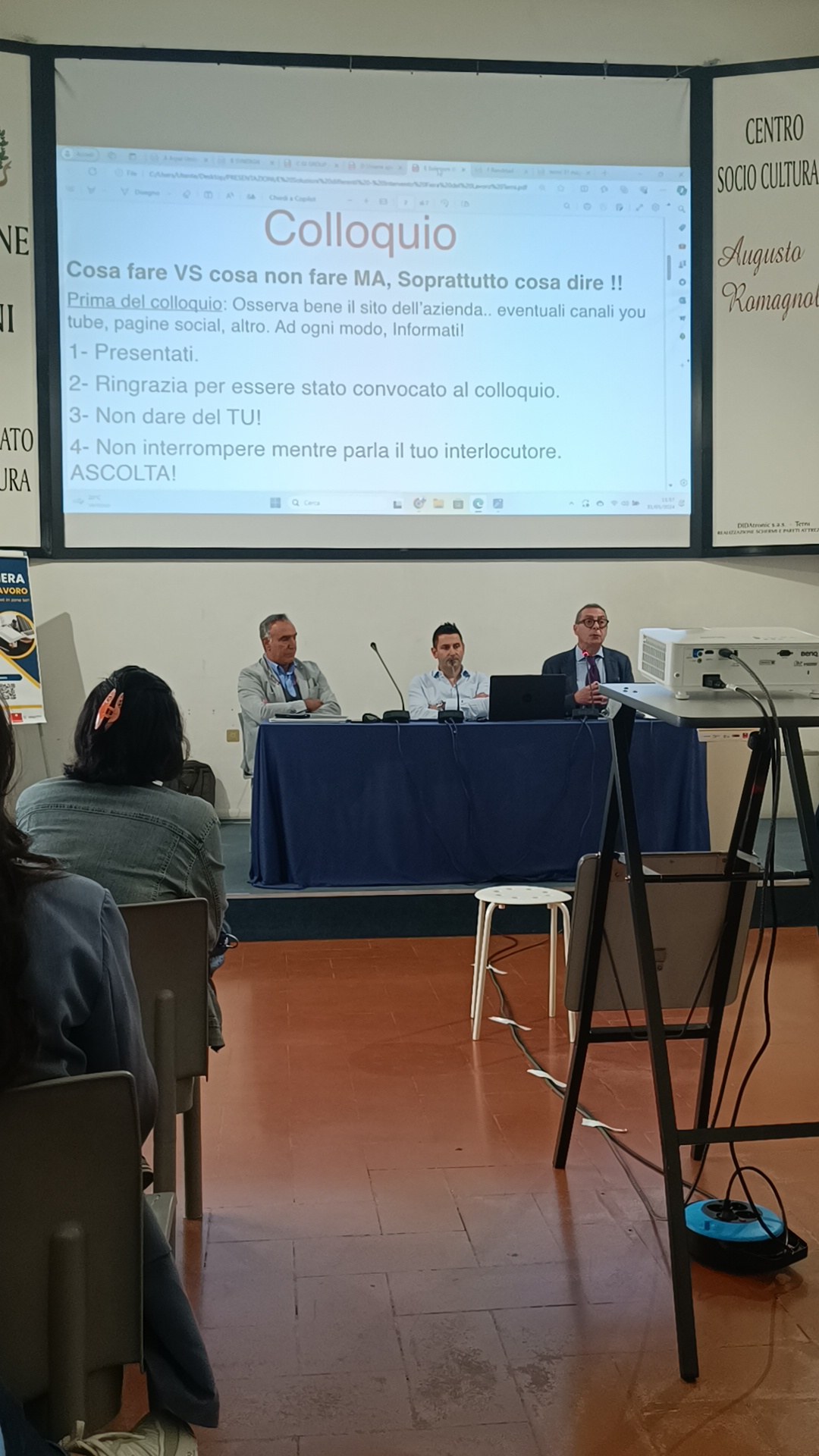 Tre relatori a un tavolo blu sotto la slide «Colloquio» davanti al pubblico, Centro Socio Culturale Augusto Romagnoli