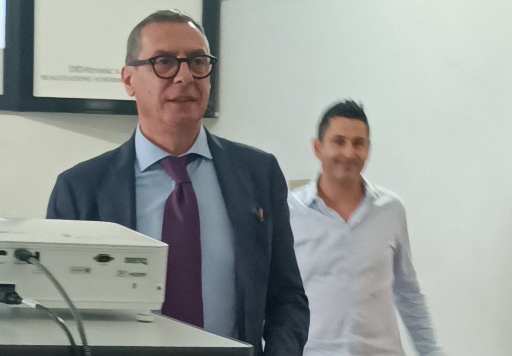 Uomo con occhiali scuri, giacca blu e cravatta viola accanto a un proiettore BenQ, alle spalle un collega in camicia chiara