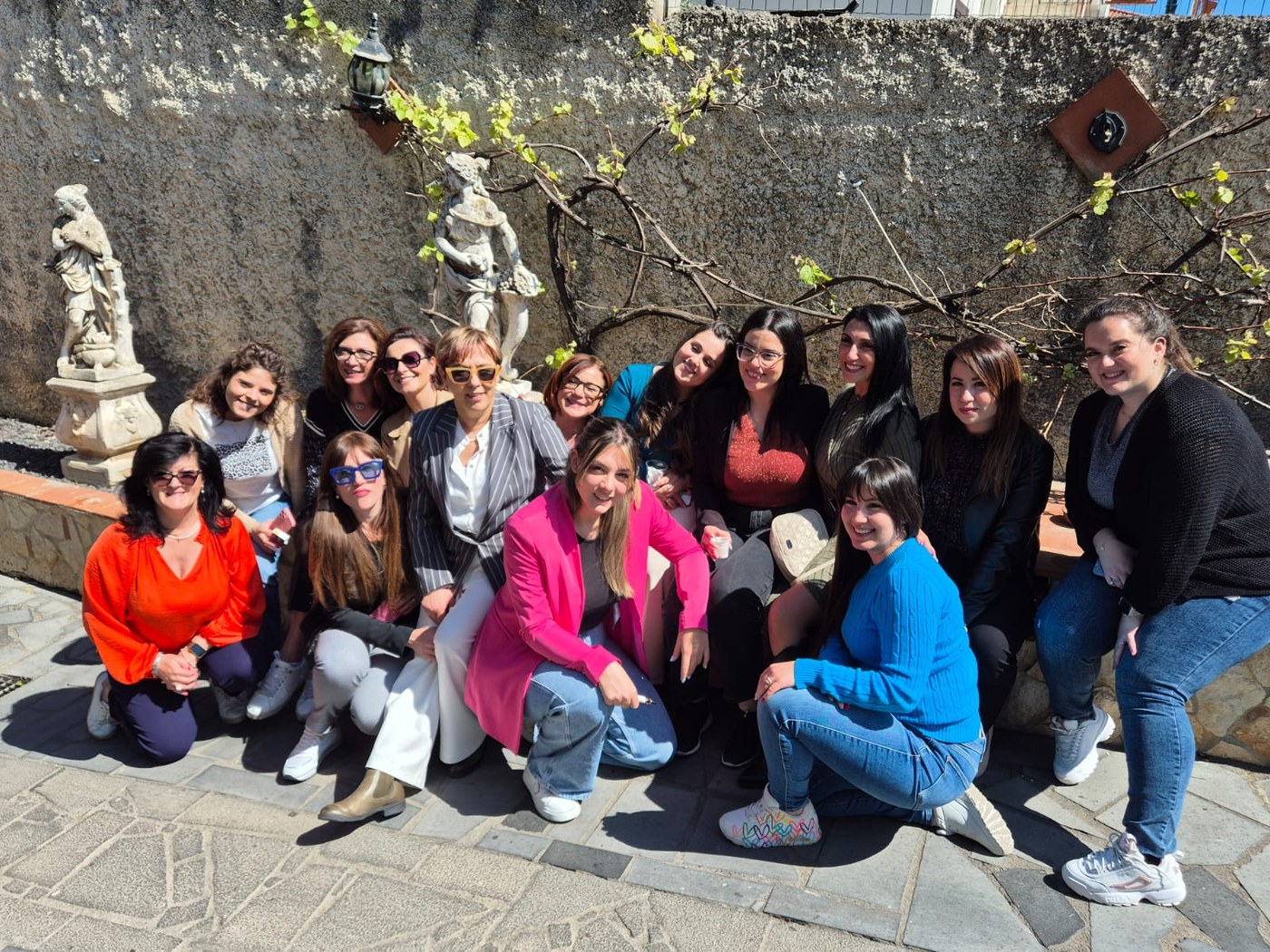 Gruppo sorridente di tredici donne in posa al sole davanti a un muro di pietra con statue e una vite